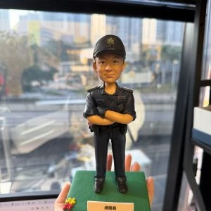 CUTEFIGUREHK 客製化香港海關人員人像公仔。公仔生動還原了人物的面貌，並穿著整齊的深藍色海關制服與海關帽，雙手交叉胸前，展現專業自信的姿態。公仔站立於點綴了小花的綠色底座上，前方設有印著「總關員 黃文傑 榮休之喜」的專屬紀念牌。這是一份專為紀律部隊人員度身訂造的退休致敬禮物。