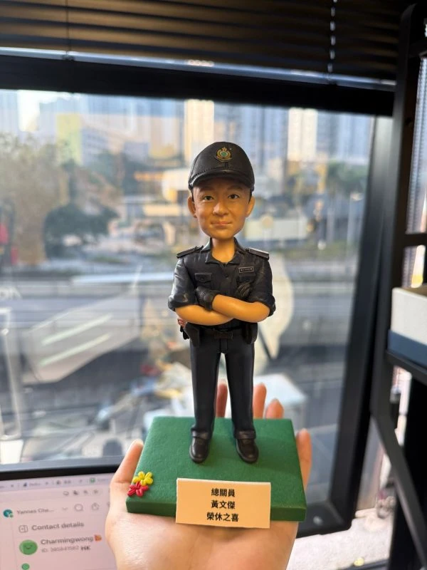 CUTEFIGUREHK 客製化香港海關人員人像公仔。公仔生動還原了人物的面貌，並穿著整齊的深藍色海關制服與海關帽，雙手交叉胸前，展現專業自信的姿態。公仔站立於點綴了小花的綠色底座上，前方設有印著「總關員 黃文傑 榮休之喜」的專屬紀念牌。這是一份專為紀律部隊人員度身訂造的退休致敬禮物。