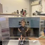 CUTEFIGUREHK 客製化榮休紀念公仔。一位戴著眼鏡的男士，身穿黑色運動套裝（上衣印有心形圖案及數字「168」，短褲亦印有「168」），雙手叉腰，右腳踩著一個足球，站立在仿石磚紋理的灰色底座上。底座前方設有一塊專屬名牌，印有「赤柱監獄高級文書主任 李永祥先生 榮休之喜」。公仔背後配有一幅印有香港懲教署徽章及「赤柱監獄 Stanley Prison」字樣的建築物背景圖，整體安放在一個透明的亞加力膠展示盒內。