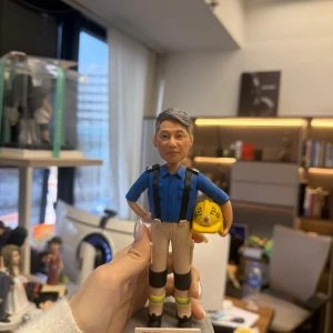 CUTEFIGUREHK 客製化榮休紀念公仔。一位灰髮男士,身穿香港消防處藍色工作服、黑色吊帶、配有護膝及反光條的卡其色長褲。他右手叉腰,左手抱著一頂印有消防徽章及「TST」字樣的黃色消防頭盔。公仔站立在仿石磚紋理的圓形灰色底座上。底座前方設有一塊專屬名牌,印有「消防總隊目 PFn11407 曾紹斌 敬業樂業33年 榮休快樂」。