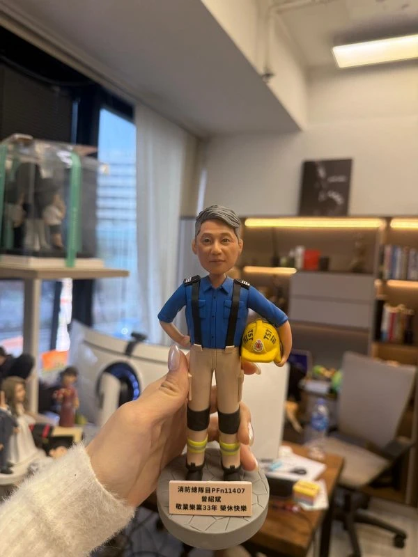 CUTEFIGUREHK 客製化榮休紀念公仔。一位灰髮男士，身穿香港消防處藍色工作服、黑色吊帶、配有護膝及反光條的卡其色長褲。他右手叉腰，左手抱著一頂印有消防徽章及「TST」字樣的黃色消防頭盔。公仔站立在仿石磚紋理的圓形灰色底座上。底座前方設有一塊專屬名牌，印有「消防總隊目 PFn11407 曾紹斌 敬業樂業33年 榮休快樂」。