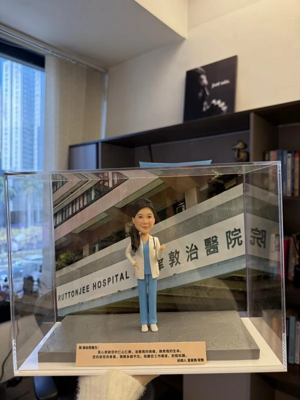 CUTEFIGUREHK 客製化女性醫生人像公仔，身穿藍色手術服和白袍並掛有聽診器。公仔放置於透明展示盒內，背景為律敦治醫院照片，底座設有刻上「仁心仁術」等真摯感謝字句的專屬牌匾，是病人送給醫生的獨一無二答謝禮物。