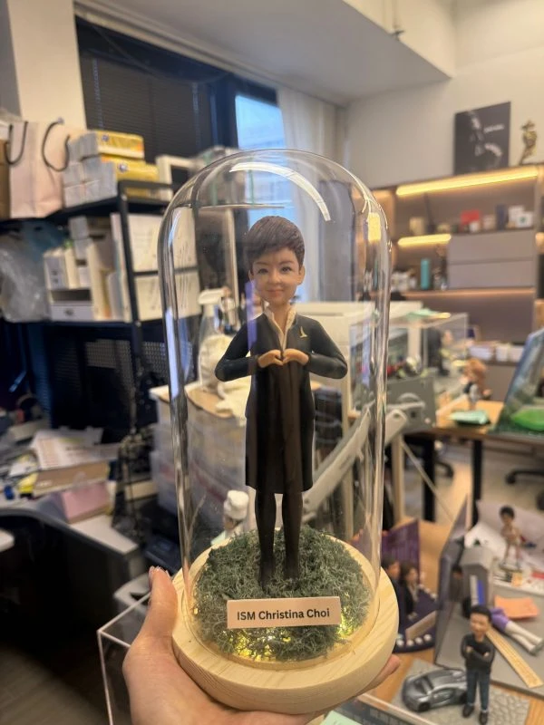 CUTEFIGUREHK 客製化空服員制服公仔。短髮造型，身穿深色空姐制服配戴金色徽章，雙手比出愛心手勢。公仔放置於帶有仿真草地及LED燈串的木底座玻璃罩內，前方設有印著「ISM Christina Choi」的專屬名牌，是送給航空業人士的升職或榮休紀念禮物。