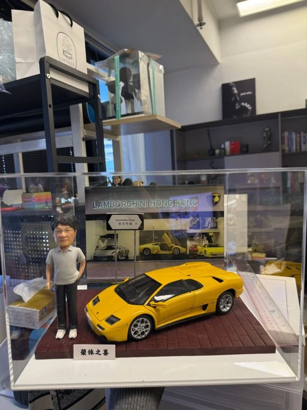 CUTEFIGUREHK 客製化人像公仔連跑車場景模型。透明展示盒內，一位穿著灰色Polo衫的男士公仔站在一輛黃色林寶堅尼跑車模型旁。背景設有「LAMBORGHINI HONG KONG」字樣的圖片背板及「告士打道」迷你路牌，底座前方印有「榮休之喜」銘牌。這是一份結合個人愛好與職業背景的專屬退休禮物。
