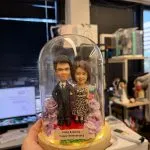 CUTEFIGUREHK 客製化情侶真人像公仔，精心佈置於帶有 LED 燈串的木座發光玻璃罩內。公仔造型為穿著西裝的男士與穿著豹紋洋裝的女士，周圍點綴著浪漫的紫色與粉色玫瑰花。底座名牌刻有「Ricky & Gloria Happy Anniversary」，是一份專為情侶或夫妻打造的浪漫週年紀念禮物。