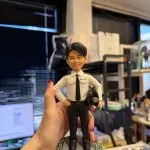 CUTEFIGUREHK 客製化男性警察人像公仔。公仔身穿香港警察經典白色制服襯衫配黑色領帶，胸前印有「POLICE 警察」字樣，左手拿著警帽，右手叉腰，展現自信英姿。公仔站立於帶有綠草與粉白小花點綴的灰色小徑圓形底座上，是一份精緻且極具紀念價值的紀律部隊禮物。