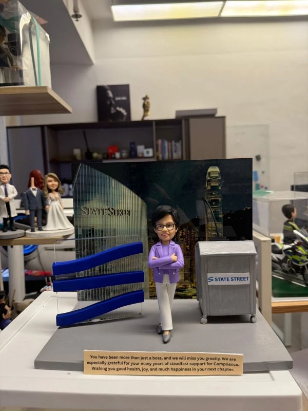 CUTEFIGUREHK 客製化企業高管紀念公仔。一位戴著黑框眼鏡的短髮女士，身穿優雅的紫色西裝外套和白色長褲，雙手交叉抱胸，展現自信專業的姿態。她站在一個精心設計的場景中，背景是印有「STATE STREET」標誌的香港中環夜景照片。場景左側有三個藍色的波浪形立體裝飾，右側有一個印有「STATE STREET」標誌的灰色演講台。底座前方有一塊刻有英文感謝語的長方形牌子，表達對這位上司多年來支持的感激與祝福。