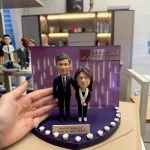CUTEFIGUREHK 客製化雙人感恩紀念公仔。一男一女穿著專業西裝與套裝的公仔，站在以香港中文大學 (CUHK) 為背景的紫色展示台上。深紫色底座邊緣裝飾著白玫瑰，並設有寫著「Heartfelt thanks for being our great leader!」的專屬名牌，是送給上司或教授的精美告別禮物。