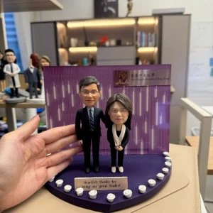 CUTEFIGUREHK 客製化雙人感恩紀念公仔。一男一女穿著專業西裝與套裝的公仔，站在以香港中文大學 (CUHK) 為背景的紫色展示台上。深紫色底座邊緣裝飾著白玫瑰，並設有寫著「Heartfelt thanks for being our great leader!」的專屬名牌，是送給上司或教授的精美告別禮物。