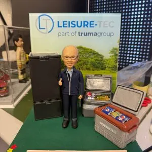 CUTEFIGUREHK 客製化企業高管及上司榮休公仔。戴眼鏡的男士身穿藍色外套比出讚好手勢，身旁配有精緻的黑色雪櫃及裝滿微縮飲品的保溫箱模型。背板印有「LEISURE-TEC」公司標誌，綠色底座前方設有寫上感謝語句的專屬名牌，是送給老闆或導師的最佳退休紀念禮物。