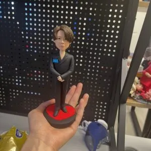 一隻手托著一個客製化單人真人公仔。公仔是一位留著棕色中長髮的男性，身穿黑色雙排扣西裝套裝與黑色皮鞋，雙手交疊在身前。他的右胸前別著一個藍色的小名牌，上面隱約寫著「Viu」。公仔站在一個圓形底座上，底座表面有一個紅色的愛心圖案。背景為一個黑色的洞洞板層架與一些雜物。
