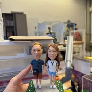 CUTEFIGUREHK 客製化情侶牽手公仔。男士穿深藍色上衣斜揹黑包，女士穿白色上衣配牛仔短褲，兩人手牽手站立於帶有綠草地與小花的灰色石板小路底座上，展現溫馨浪漫的日常散步情境，是完美的週年紀念或情人節禮物。