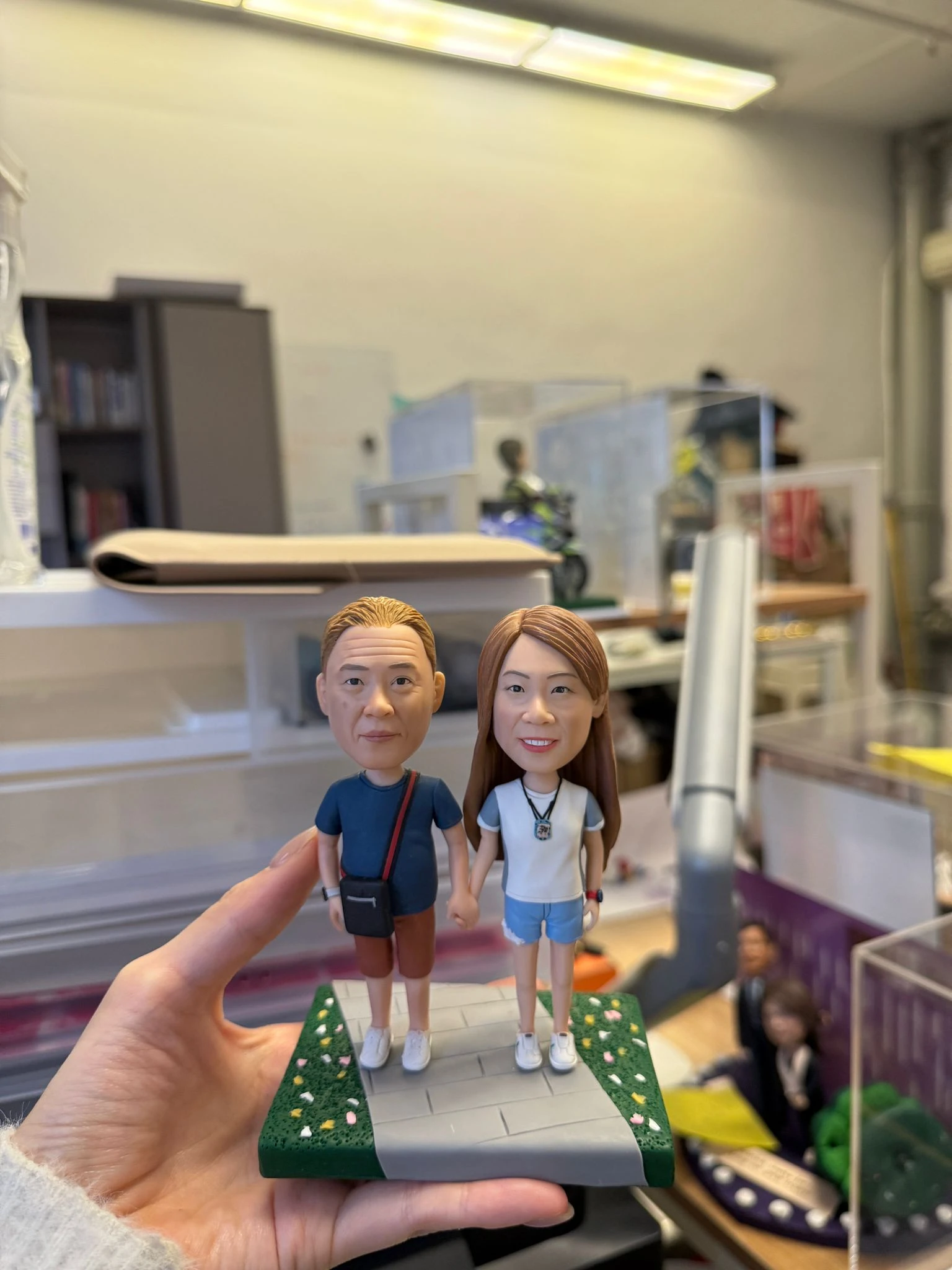 CUTEFIGUREHK 客製化情侶牽手公仔。男士穿深藍色上衣斜揹黑包，女士穿白色上衣配牛仔短褲，兩人手牽手站立於帶有綠草地與小花的灰色石板小路底座上，展現溫馨浪漫的日常散步情境，是完美的週年紀念或情人節禮物。
