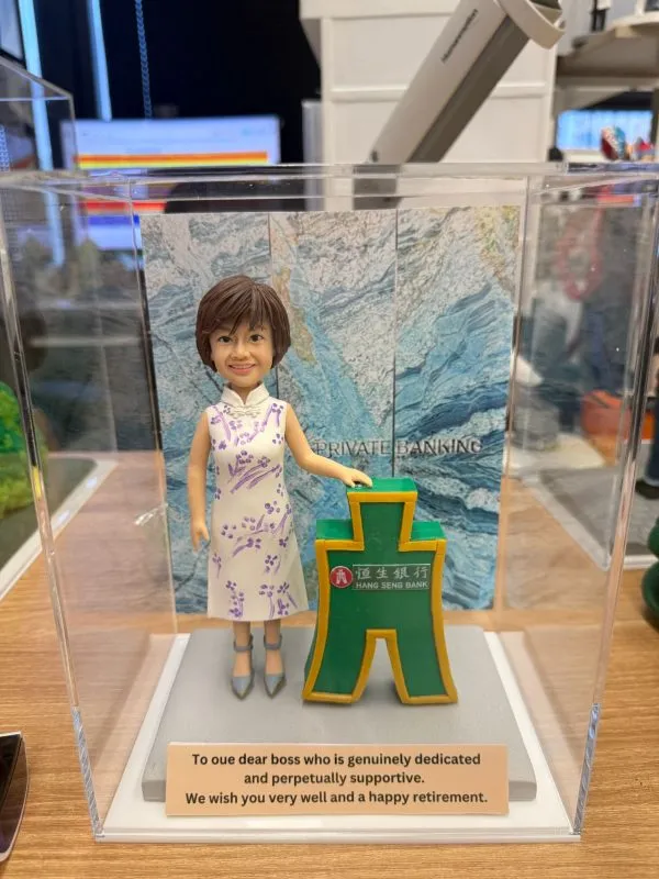 一尊裝在透明壓克力展示盒內的客製化女性公仔，作為銀行主管的退休禮物。公仔為短髮女性，笑容親切，身穿白色帶有紫色花紋的無袖旗袍。她的左手搭在一個帶有「恒生銀行 HANG SENG BANK」標誌的綠色古錢幣立體模型上。展示盒內的背景板印有藍色紋理與「PRIVATE BANKING」字樣。底座前方有一塊銘牌，以英文寫著對老闆的感謝與退休祝福。