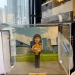 CUTEFIGUREHK 客製化退休紀念真人像公仔，裝於透明壓克力展示盒內。公仔造型為穿著黑色套裝、手持繽紛花束的短髮女士。展示盒背景為香港添馬公園與政府總部大樓風景照。底座名牌刻有「Apple Happy Retirement 2026」，是一份專為慶祝榮休而訂製的專屬紀念禮物。