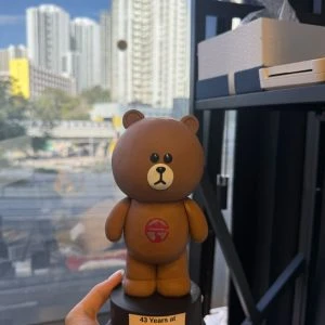 CUTEFIGUREHK 客製化紀念公仔。一個手工製作的 LINE FRIENDS 熊大(Brown)立體公仔,站立在黑色圓形底座上。熊大胸前印有一個紅色的圓形標誌(類似嶺南大學校徽)。黑色底座上貼有一張白色標籤,上面印有黑色英文字:「43 Years at Lingnan / In thy care bright / years are bringing.」。背景是透過窗戶看到的城市高樓大廈和藍天。