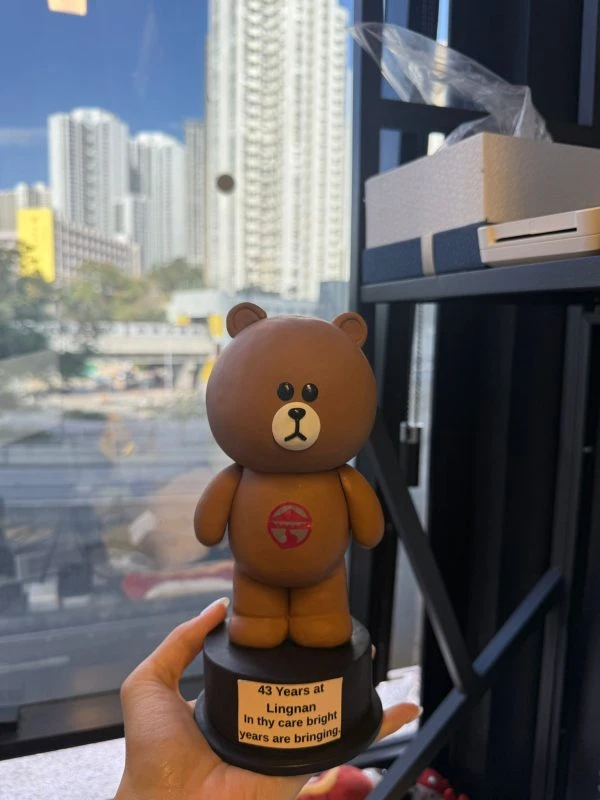CUTEFIGUREHK 客製化紀念公仔。一個手工製作的 LINE FRIENDS 熊大（Brown）立體公仔，站立在黑色圓形底座上。熊大胸前印有一個紅色的圓形標誌（類似嶺南大學校徽）。黑色底座上貼有一張白色標籤，上面印有黑色英文字：「43 Years at Lingnan / In thy care bright / years are bringing.」。背景是透過窗戶看到的城市高樓大廈和藍天。