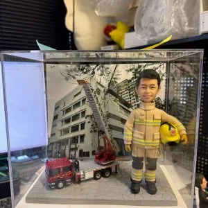 CUTEFIGUREHK 客製化消防員紀念公仔。一位身穿黃褐色消防抗火服(帶有黃色反光條紋及黑色護膝)的男士,左手抱著印有徽章的黃色消防頭盔,笑容自信。公仔放置在透明展示盒內,站在灰色石板路面設計的底座上,旁邊停泊著一輛精緻的迷你紅色消防雲梯車模型。背景板印有消防局大樓的外觀照片。這是送給消防員退休或升職的極佳紀念禮物。