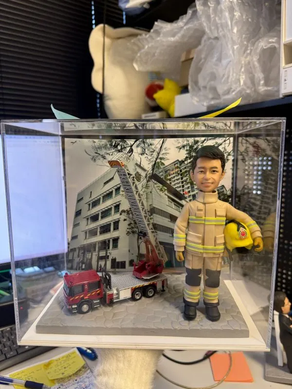 CUTEFIGUREHK 客製化消防員紀念公仔。一位身穿黃褐色消防抗火服（帶有黃色反光條紋及黑色護膝）的男士，左手抱著印有徽章的黃色消防頭盔，笑容自信。公仔放置在透明展示盒內，站在灰色石板路面設計的底座上，旁邊停泊著一輛精緻的迷你紅色消防雲梯車模型。背景板印有消防局大樓的外觀照片。這是送給消防員退休或升職的極佳紀念禮物。