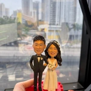 CUTEFIGUREHK 客製化結婚人像公仔，生動還原了身穿黑色西裝打領結的新郎，以及穿著白色深V婚紗、手持白色玫瑰花球的新娘。兩人甜蜜挽手，站立於帶有立體小玫瑰裝飾的精緻紅色心形底座上。這是一份極具心思的新婚送禮、婚禮迎賓桌（Reception）佈置或結婚週年紀念禮物。
