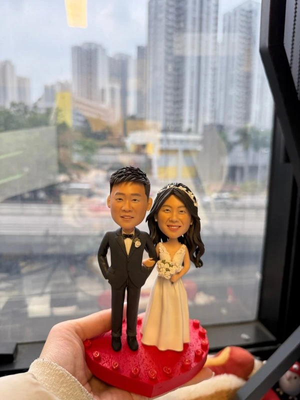 CUTEFIGUREHK 客製化結婚人像公仔，生動還原了身穿黑色西裝打領結的新郎，以及穿著白色深V婚紗、手持白色玫瑰花球的新娘。兩人甜蜜挽手，站立於帶有立體小玫瑰裝飾的精緻紅色心形底座上。這是一份極具心思的新婚送禮、婚禮迎賓桌（Reception）佈置或結婚週年紀念禮物。
