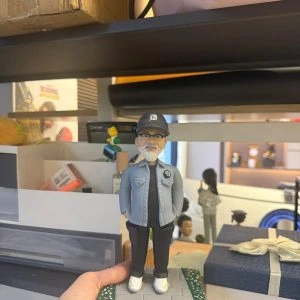 CUTEFIGUREHK 客製化男士人像公仔,生動還原了戴著黑色鴨舌帽與眼鏡、留著標誌性白鬍鬚的男士。他身穿淺藍色休閒外套、黑色上衣及工裝褲,雙手插袋,展現型格休閒風。公仔站立於帶有草地小徑的底座上,前方設有寫著「Dear David, Lululemon 10th anniversary」的專屬紀念牌。這是一份極具心思、專為慶祝員工入職十週年及長期服務而設的企業紀念禮物。