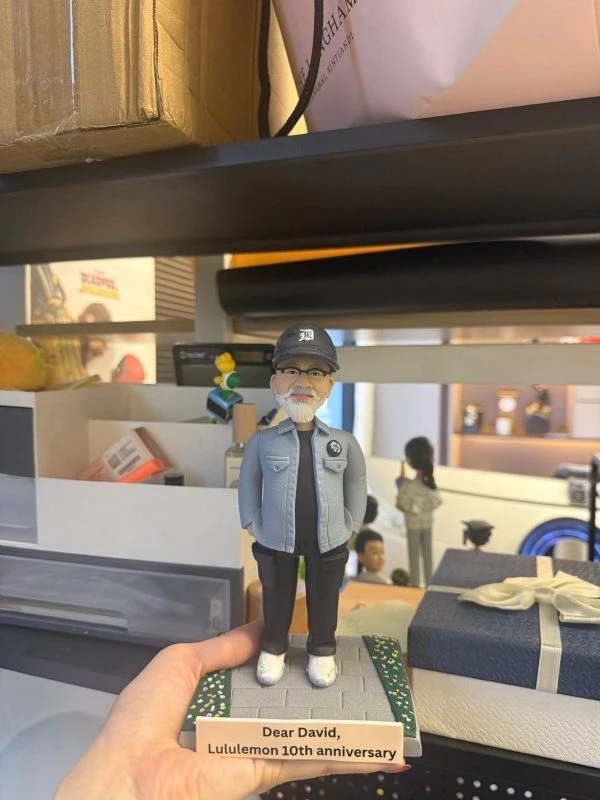 CUTEFIGUREHK 客製化男士人像公仔，生動還原了戴著黑色鴨舌帽與眼鏡、留著標誌性白鬍鬚的男士。他身穿淺藍色休閒外套、黑色上衣及工裝褲，雙手插袋，展現型格休閒風。公仔站立於帶有草地小徑的底座上，前方設有寫著「Dear David, Lululemon 10th anniversary」的專屬紀念牌。這是一份極具心思、專為慶祝員工入職十週年及長期服務而設的企業紀念禮物。