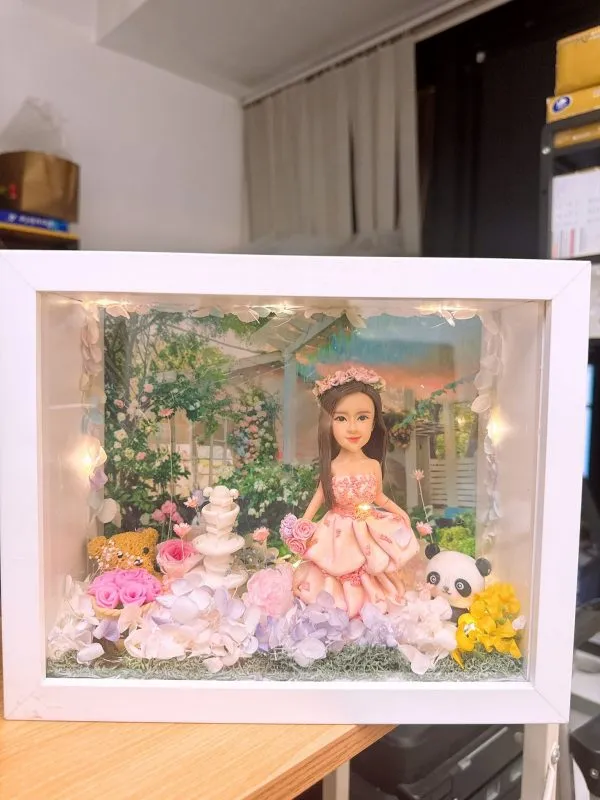 CUTEFIGUREHK 客製化立體相框公仔禮物。一個白色的發光畫框，內部佈置成夢幻的童話花園。中央是一個穿著粉紅色花瓣晚裝的長髮女孩立體公仔，頭戴花環，手持粉紅玫瑰。背景是浪漫的黃昏花園圖案，公仔腳下鋪滿了紫色、白色和粉紅色的乾花（繡球花）以及綠色苔蘚，旁邊點綴著微縮的白色小噴泉、可愛的小棕熊和熊貓模型。相框內置有溫馨的 LED 燈串，營造出唯美的氣氛。