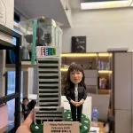 CUTEFIGUREHK 客製化榮休紀念公仔。一位穿著黑色套裝、佩戴員工證的女職員公仔，站在帶有 FEHD 標誌的微縮大樓模型旁。綠色底座點綴著花瓣，並設有寫著「Happy Retirement February 2026 身體健康 享樂人生」的專屬名牌，是專為食環署職員訂製的精美退休禮物。