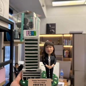CUTEFIGUREHK 客製化榮休紀念公仔。一位穿著黑色套裝、佩戴員工證的女職員公仔，站在帶有 FEHD 標誌的微縮大樓模型旁。綠色底座點綴著花瓣，並設有寫著「Happy Retirement February 2026 身體健康 享樂人生」的專屬名牌，是專為食環署職員訂製的精美退休禮物。