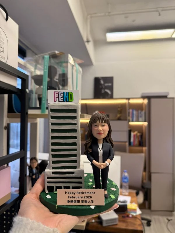 CUTEFIGUREHK 客製化榮休紀念公仔。一位穿著黑色套裝、佩戴員工證的女職員公仔，站在帶有 FEHD 標誌的微縮大樓模型旁。綠色底座點綴著花瓣，並設有寫著「Happy Retirement February 2026 身體健康 享樂人生」的專屬名牌，是專為食環署職員訂製的精美退休禮物。