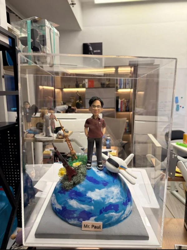 CUTEFIGUREHK 客製化環遊世界主題紀念公仔。一位戴眼鏡的男士身穿酒紅色短袖Polo衫和黑色長褲，雙手插袋。公仔站立在一個藍白相間、仿如地球表面的半球體底座上。底座上裝飾有一架白色飛機模型，以及巴黎鐵塔和東方明珠塔的微縮模型，並點綴著綠色苔蘚和黃色小花。底座前方設有一塊名牌，印有「Mr. Paul」。整個模型被放置在透明展示盒內。