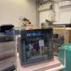 CUTEFIGUREHK 客製化店舖微縮模型。一個精緻的微型髮型屋店面模型，頂部有黑底藍字及白字的招牌寫著「Modi Hair」和「安寧路49號」。店面採用透明玻璃門設計，內部貼有價目表海報，隱約可見店內的裝潢和一張黃色高腳椅。模型放置在一個淺粉紅色的方形底座上，並罩著透明的壓克力展示盒。背景是隔著玻璃窗看到的城市高樓大廈和藍天。