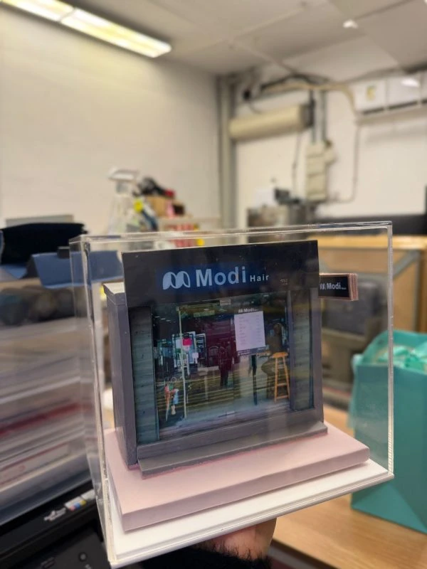 CUTEFIGUREHK 客製化店舖微縮模型。一個精緻的微型髮型屋店面模型，頂部有黑底藍字及白字的招牌寫著「Modi Hair」和「安寧路49號」。店面採用透明玻璃門設計，內部貼有價目表海報，隱約可見店內的裝潢和一張黃色高腳椅。模型放置在一個淺粉紅色的方形底座上，並罩著透明的壓克力展示盒。背景是隔著玻璃窗看到的城市高樓大廈和藍天。