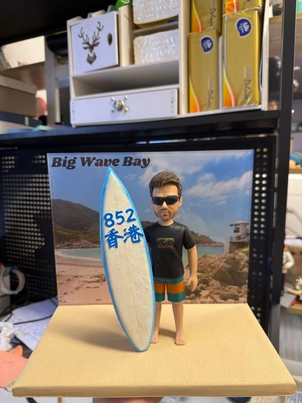 客製化單人衝浪主題真人公仔。公仔是一位留著棕色短髮與落腮鬍的男性，戴著黑色墨鏡，身穿印有波浪圖案的黑色短袖T恤與藍橘相間的衝浪褲，赤腳站立。他右手扶著一塊白底藍邊的衝浪板，板上寫有藍色的「852」與「香港」字樣。公仔站在模擬沙灘的淺黃色方形底座上，背後有一張印有「Big Wave Bay」字樣及海灘風景（包含沙灘、海浪、山丘與救生塔）的背景板。背景為室內層架與雜物。