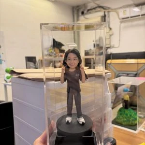 一尊裝在透明壓克力展示盒內的客製化女性公仔。公仔為長髮女性，笑容燦爛，雙手比出「YA」的手勢。她身穿深灰色短袖上衣（左胸印有「趙」字）與同色系長褲，腳穿白色厚底休閒鞋。公仔站在黑色圓形底座上，底座前方印有金色的「The Best Mum」字樣。背景為工作室環境。