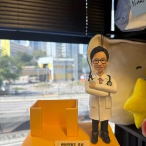 CUTEFIGUREHK 客製化醫生禮物公仔。一個穿著白袍、戴著黑框眼鏡和聽診器的男醫生立體公仔，雙手交叉抱胸，展現專業自信的笑容。公仔站在方形黃色底座上，旁邊配有一個實用的黃色名片座（或筆筒）設計。底座前方鑲有一塊客製化銘牌，上面印有「黃劍明醫生 惠存 仁心仁術 方松洲家人 敬贈」的致謝字句。背景為窗外的城市街景。
