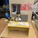 CUTEFIGUREHK 客製化退休禮物公仔。一個精緻的辦公室場景立體模型，主角是一位短髮女士，身穿黑白格紋外套，雙手交叉自信地坐在辦公桌前。黃色的辦公桌上放滿了文件和一部白色電話。旁邊的側桌有一部顯示 Windows 11 畫面的電腦屏幕及鍵盤滑鼠。背景牆上有窗戶透出城市高樓景色，窗台放有打印機，牆上貼有「2025 香港義工獎」及「國際義工日」的縮微海報。模型前方底座有一塊紅色名牌，用白字寫著「Goodbye documents, Hello Husband」及「Likie Lee CEO」。