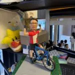 客製化單人自行車真人公仔。公仔是一位戴著黑框眼鏡的男性，身穿紅底白塊的短袖上衣、黑色單車褲與黑色半指手套。他跨騎在一輛藍色公路自行車上，雙手握拳高舉，擺出充滿活力與自信的慶祝姿勢。公仔固定在一個模擬灰色柏油路與兩側綠色草地（點綴著彩色小花）的方形底座上。底座前方有一塊白色字牌，上面印有中文字：「KP, 祝願你開啟新篇章，享受慢活，悠遊自在的生活。」。背景為擺放雜物的層架。