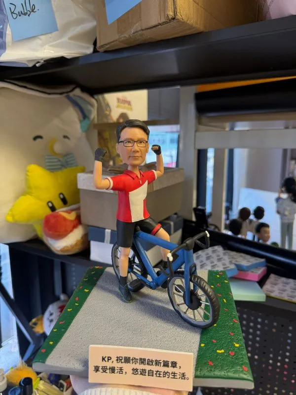客製化單人自行車真人公仔。公仔是一位戴著黑框眼鏡的男性，身穿紅底白塊的短袖上衣、黑色單車褲與黑色半指手套。他跨騎在一輛藍色公路自行車上，雙手握拳高舉，擺出充滿活力與自信的慶祝姿勢。公仔固定在一個模擬灰色柏油路與兩側綠色草地（點綴著彩色小花）的方形底座上。底座前方有一塊白色字牌，上面印有中文字：「KP, 祝願你開啟新篇章，享受慢活，悠遊自在的生活。」。背景為擺放雜物的層架。
