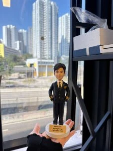 CUTEFIGUREHK 客製化飛機師禮物公仔。一隻手托著一個精緻的立體真人公仔，背景是城市高樓與公路的窗外日景。公仔主角是一位身穿深藍色飛機師制服的男士，制服袖口有金色條紋，內搭白襯衫與圖案領帶，雙手插袋，神態自信從容。公仔站在白色的圓形底座上，底座前方有一塊黃色名牌，上面用黑字寫著英文勵志金句「Never quit. Never give up. Fly it to the end.」。
