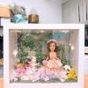 CUTEFIGUREHK 客製化立體相框公仔禮物。一個白色的發光畫框，內部佈置成夢幻的童話花園。中央是一個穿著粉紅色花瓣晚裝的長髮女孩立體公仔，頭戴花環，手持粉紅玫瑰。背景是浪漫的黃昏花園圖案，公仔腳下鋪滿了紫色、白色和粉紅色的乾花（繡球花）以及綠色苔蘚，旁邊點綴著微縮的白色小噴泉、可愛的小棕熊和熊貓模型。相框內置有溫馨的 LED 燈串，營造出唯美的氣氛。