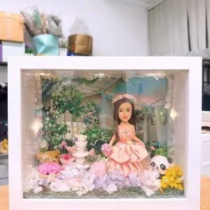 CUTEFIGUREHK 客製化立體相框公仔禮物。一個白色的發光畫框,內部佈置成夢幻的童話花園。中央是一個穿著粉紅色花瓣晚裝的長髮女孩立體公仔,頭戴花環,手持粉紅玫瑰。背景是浪漫的黃昏花園圖案,公仔腳下鋪滿了紫色、白色和粉紅色的乾花(繡球花)以及綠色苔蘚,旁邊點綴著微縮的白色小噴泉、可愛的小棕熊和熊貓模型。相框內置有溫馨的 LED 燈串,營造出唯美的氣氛。