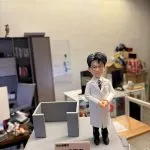 一尊客製化的男醫生立體公仔。公仔戴著黑框眼鏡，身穿白色醫師袍、打著條紋領帶與黑長褲，雙手交疊放在身前，展現專業沉穩的形象。公仔站在灰色的方形底座上，旁邊有一個灰色的簡約ㄇ字型結構。底座前方有一塊粉色名牌，上面印有「何文傑醫生」、「妙手回春」以及「張生敬贈」的字樣。背景為一個辦公室或工作室環境。