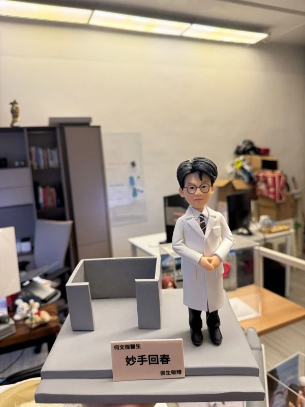 一尊客製化的男醫生立體公仔。公仔戴著黑框眼鏡，身穿白色醫師袍、打著條紋領帶與黑長褲，雙手交疊放在身前，展現專業沉穩的形象。公仔站在灰色的方形底座上，旁邊有一個灰色的簡約ㄇ字型結構。底座前方有一塊粉色名牌，上面印有「何文傑醫生」、「妙手回春」以及「張生敬贈」的字樣。背景為一個辦公室或工作室環境。