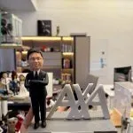 CUTEFIGUREHK 客製化男士退休公仔。公仔身穿筆挺黑色西裝、戴眼鏡並雙手交叉胸前，展現專業自信。旁邊配有大型立體 AXA 企業標誌，灰色仿石紋底座前方印有「Happy Retirement to Samuel」專屬祝賀語名牌，是完美的企業榮休紀念禮物。