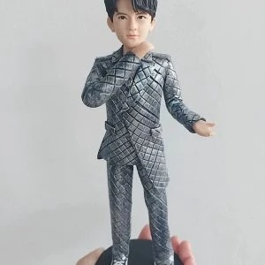 客製化單人立體真人公仔。一隻手托著一個黑色圓形底座的男性公仔。公仔有著黑色的旁分短髮,五官清秀。他穿著一套引人注目的銀灰色菱格紋西裝套裝,搭配黑白相間的休閒帆布鞋。公仔右手輕觸下巴,左手向外微攤,擺出自信且充滿魅力的姿勢。背景為純白牆面。