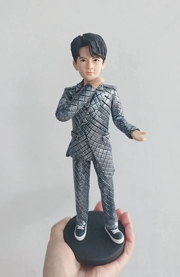 客製化單人立體真人公仔。一隻手托著一個黑色圓形底座的男性公仔。公仔有著黑色的旁分短髮，五官清秀。他穿著一套引人注目的銀灰色菱格紋西裝套裝，搭配黑白相間的休閒帆布鞋。公仔右手輕觸下巴，左手向外微攤，擺出自信且充滿魅力的姿勢。背景為純白牆面。