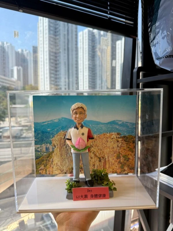 CUTEFIGUREHK 客製化長輩生日公仔。透明亞加力膠盒內展示著一個根據真人相貌製作的立體公仔。公仔是一位白髮、戴著眼鏡、笑容和藹的男士，身穿休閒 Polo 恤和牛仔褲，雙手捧著一個象徵長壽的粉紅色大壽桃。底座點綴了仿真的綠色植物，前方放有一塊紅色名牌，上面印有黑字：「Dee / 60大壽 身體健康」。展示盒內的背景是一張壯麗的高山風景照（類似香港獅子山）。