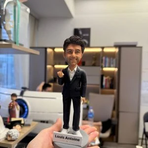 CUTEFIGUREHK 客製化男士公仔。人偶穿著深色西裝外套配白襯衫與白球鞋，展現商務休閒風格。右手舉起食指，面帶自信微笑。灰色圓形底座上帶有「Louis Antoine」專屬名牌及小金星圖案，是完美的升職或職場紀念禮物。